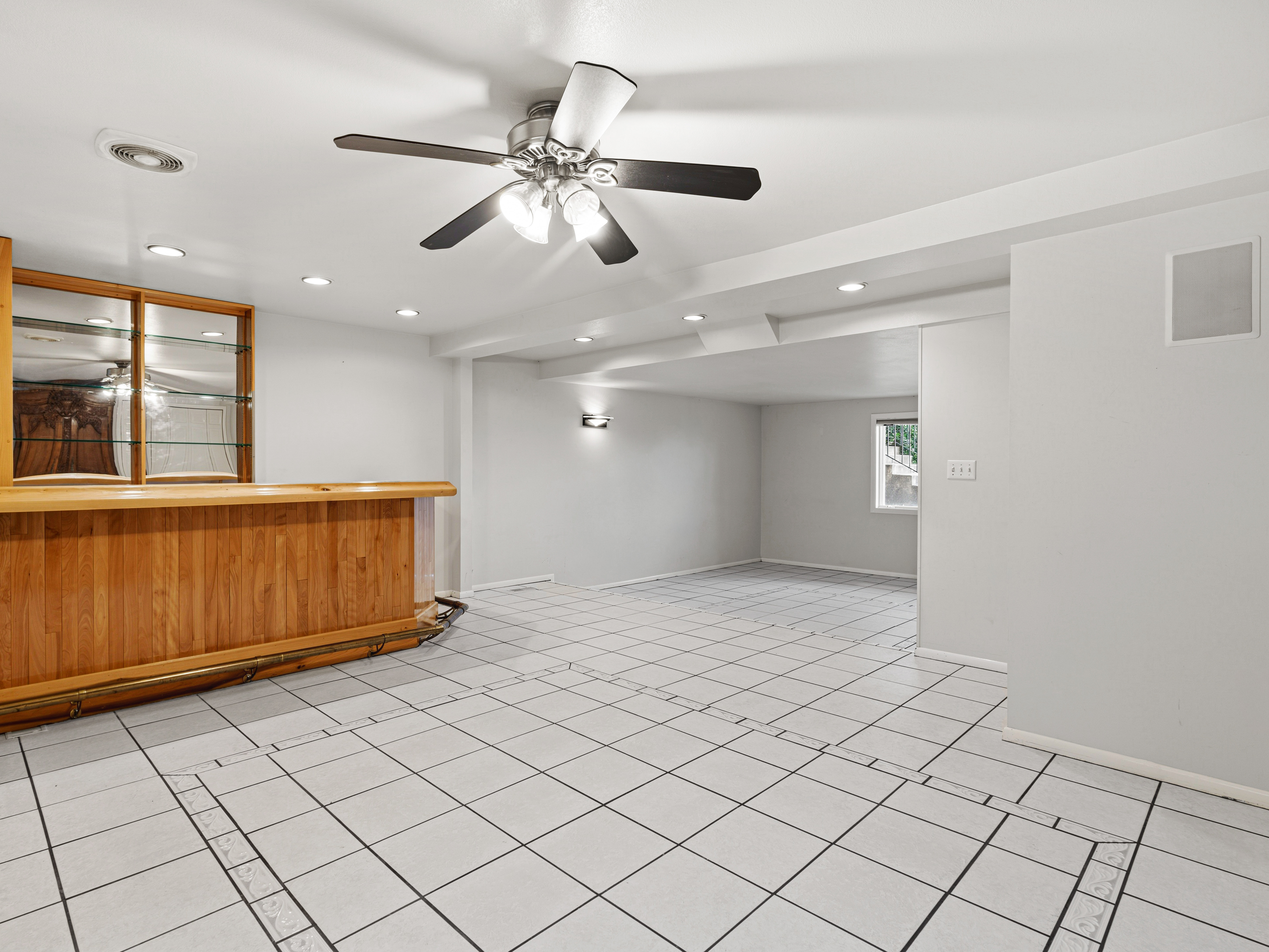 Spacious open room featuring a built-in wood bar with mirrored shelving, white tiled flooring, recessed lighting, and a ceiling fan