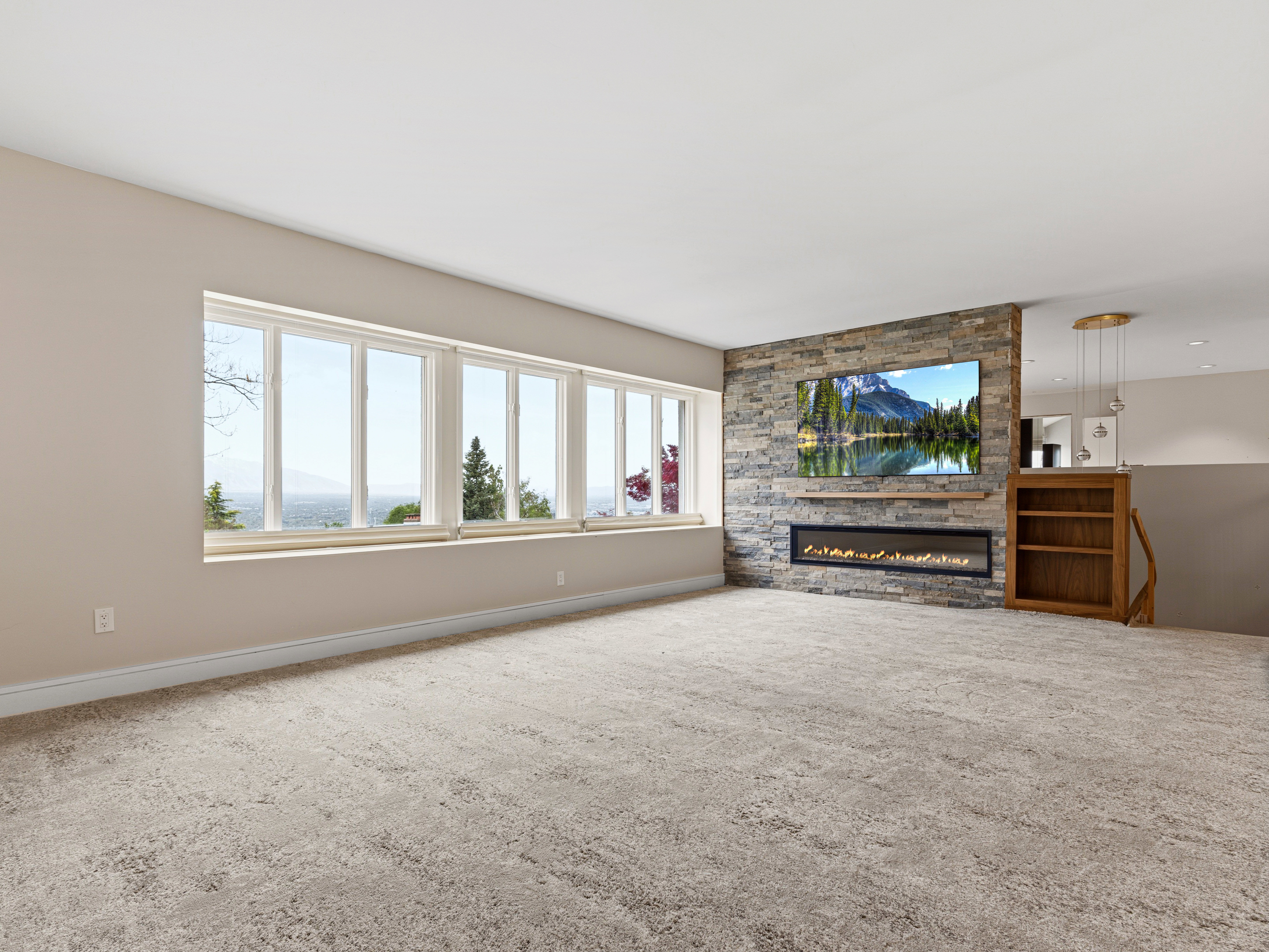 Spacious room featuring a long horizontal window, a stacked stone accent wall, an electric fireplace with a floating wood mantle, light-colored carpeting, and a built-in wooden bookcase