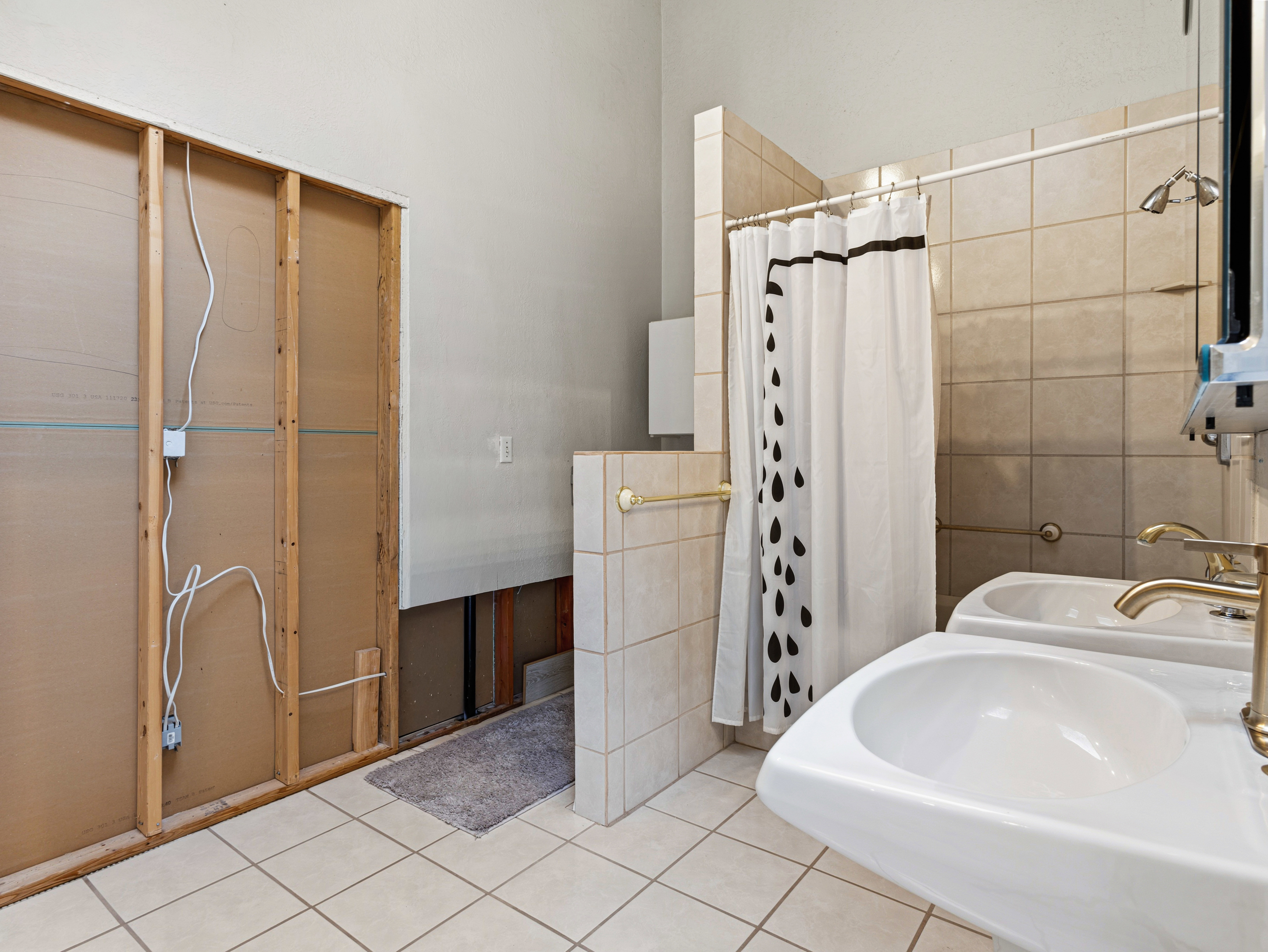 Two wall-mounted pedestal sinks with brass-finish faucets, a tiled shower area, and light-toned floor tiling