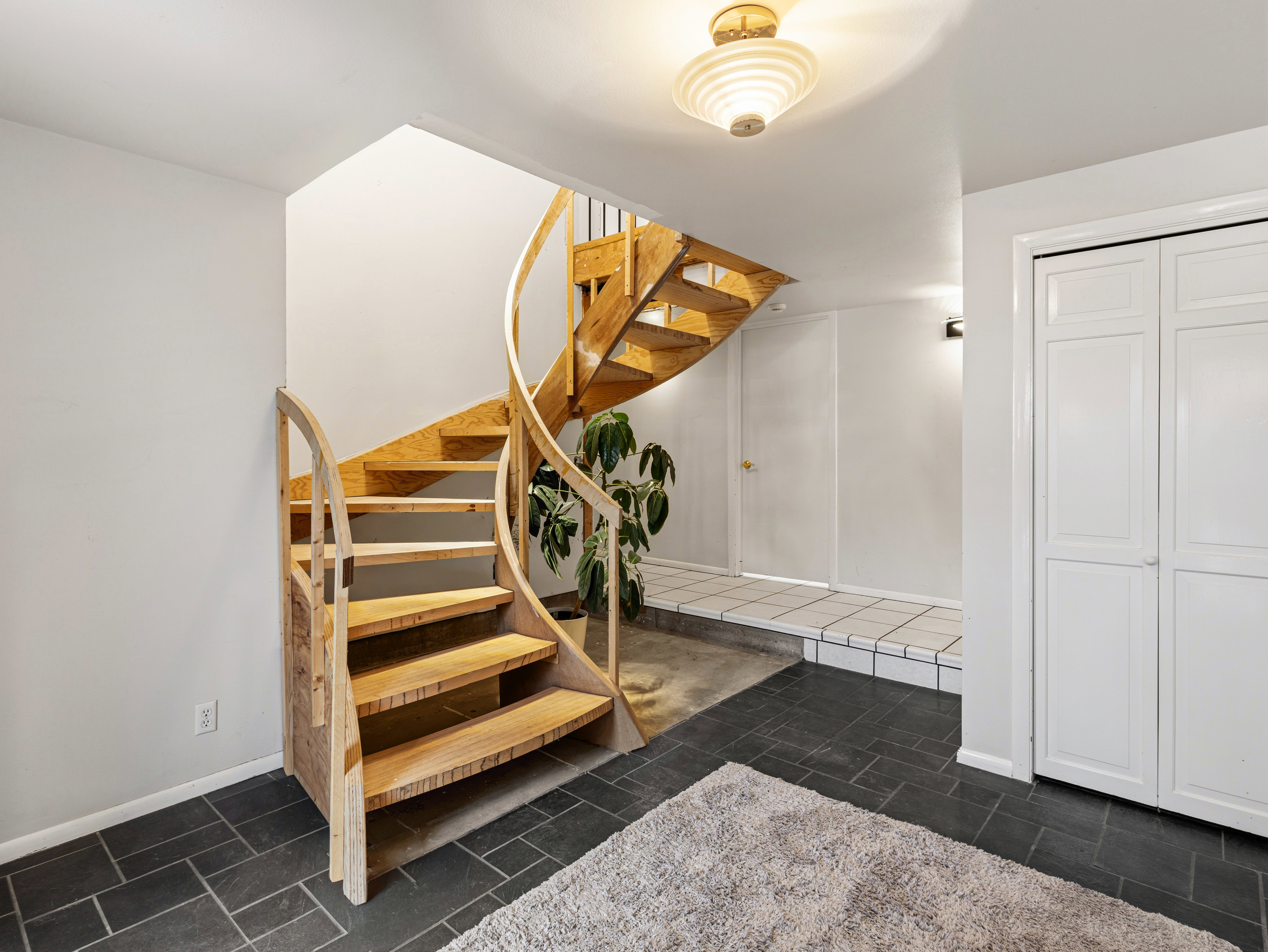Spiral staircase with natural wood treads and balusters, dark slate-style floor tiling, white walls, built-in bi-fold closet doors, and an overhead semi-flush mount light fixture