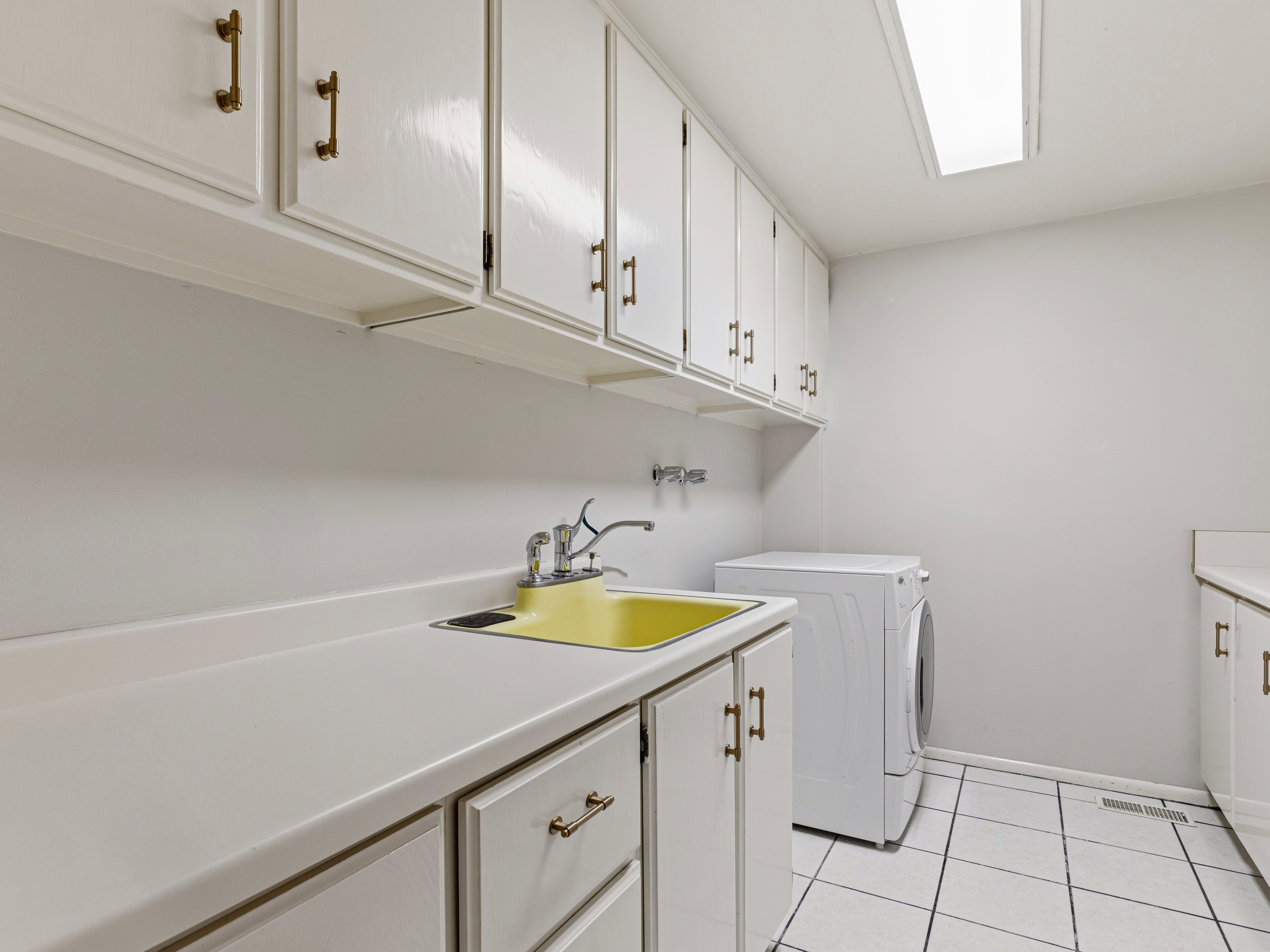 Utility area featuring white cabinetry with brass-toned hardware, extensive countertop space, a yellow utility sink with faucet, and white tiled flooring