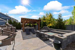 Multi-level hardscaped patio featuring a timber pergola, built-in outdoor kitchen, paver stone surfaces, tiered block wall seating, and an integrated fire pit
