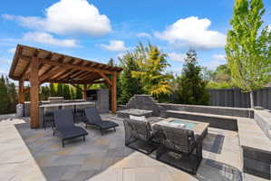 Expansive paver patio featuring a wood pergola, built-in outdoor kitchen with grill, integrated seating, and a gas fire pit