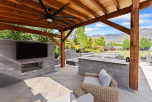 Covered outdoor patio featuring a wood beam pergola, ceiling fan, and stone-block entertainment wall with a built-in fire feature
