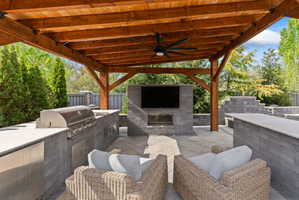 Outdoor living area featuring a wood-beam pergola with a ceiling fan, a built-in grill, stone countertops, and a mounted television above a fireplace