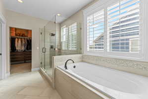 Soaking tub with a matte black faucet, glass-enclosed shower, plantation shutters, and a custom walk-in closet