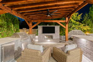 Outdoor covered patio featuring a wood beam ceiling, built-in stainless steel grill, stone block outdoor kitchen with integrated appliances, stone block media wall, and an outdoor ceiling fan