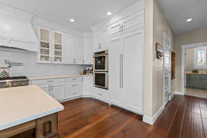 Bright kitchen featuring white cabinetry with glass-front accents, white subway tile backsplash, and stainless steel appliances including a built-in double oven and gas range with pot filler