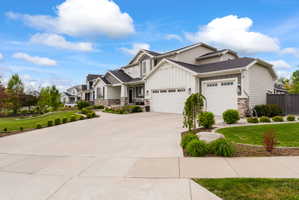 Modern colonial-style residence featuring a multi-car garage, board and batten siding accents, stone veneer detailing, a concrete driveway, and established landscape plantings