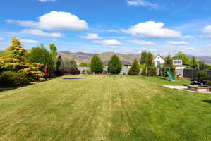 Expansive private backyard featuring a large lawn, mature trees and shrubs, a dark privacy fence, and a concrete patio with a built-in fire pit