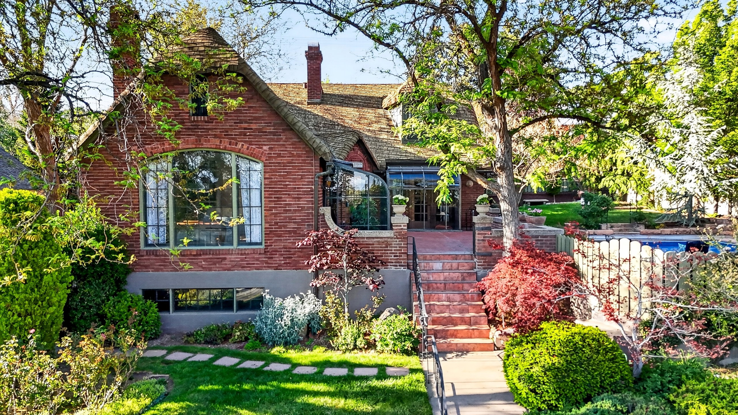 Brick exterior featuring an arched picture window, a sunroom with multiple windows, a multi-level patio with steps, and a fenced swimming pool