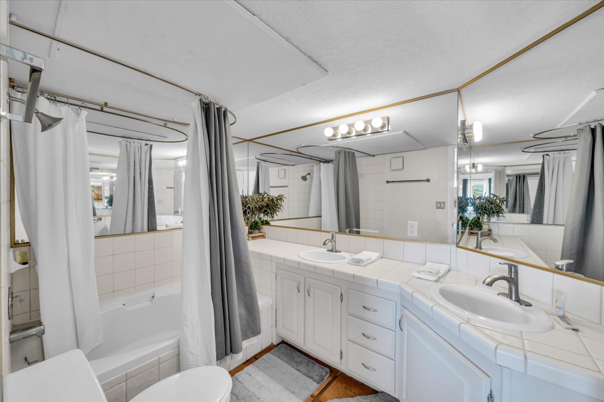 Bathroom featuring a built-in tub with white tile surround, a dual vanity with white tile countertops and integrated sinks, extensive wall-mounted mirrors, and white cabinetry with brushed nickel hardware