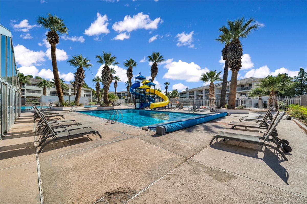 Outdoor swimming pool featuring a bright blue water slide, a concrete deck, multiple lounge chairs, and surrounding palm trees