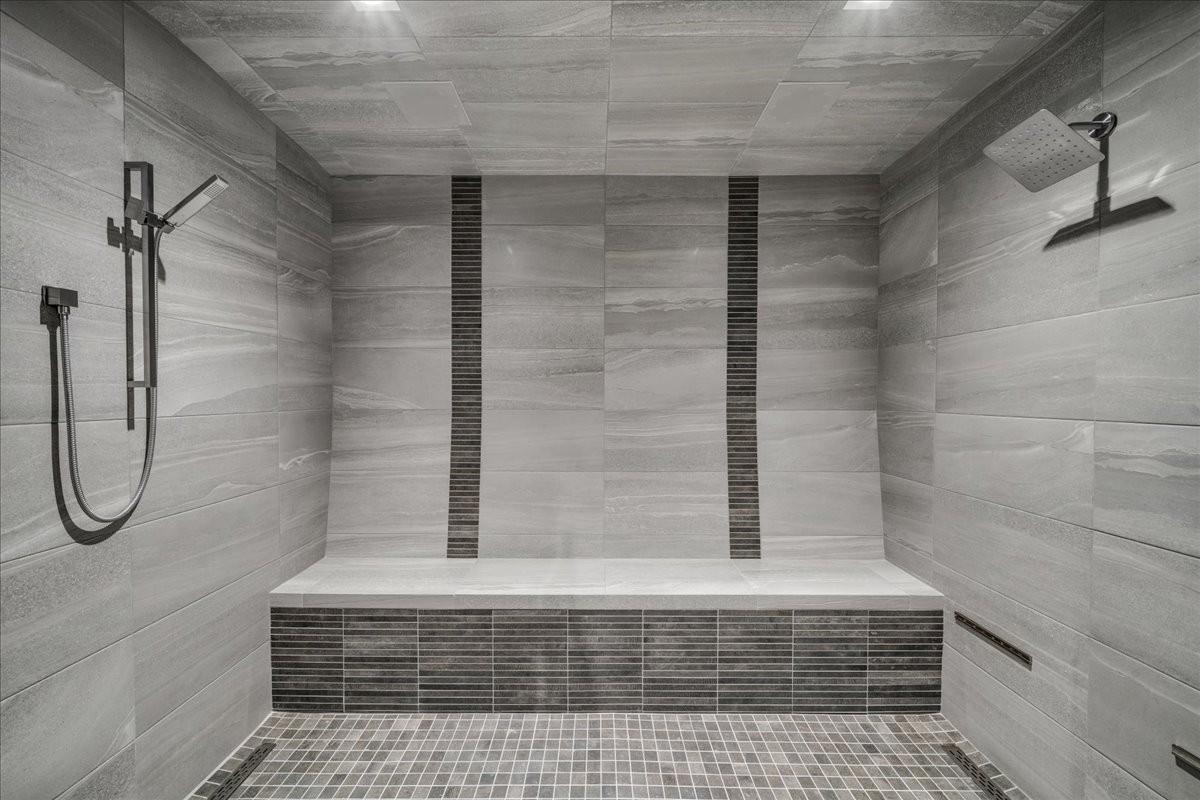 Spacious shower featuring full-height stone-look tile, a built-in bench, dual showerheads with a handheld sprayer, and mosaic tile accents
