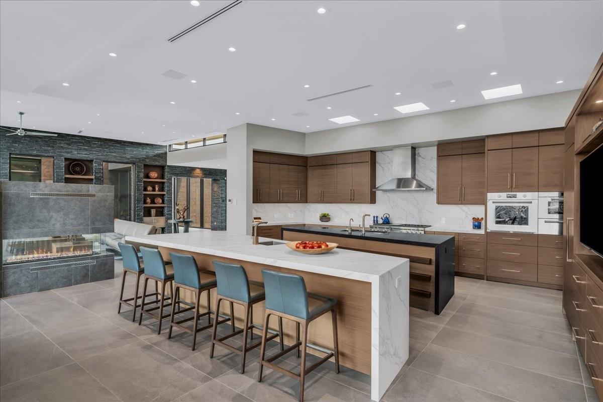 Spacious open-concept kitchen and living area featuring a large center island with waterfall countertop, a second island with integrated sink, wood-finish cabinetry, and a linear fireplace with stone surround