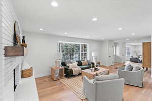 Spacious living area featuring light wood-finish flooring, recessed lighting, and a white brick fireplace with a floating wood mantle