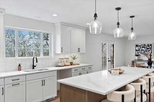 Contemporary kitchen featuring white cabinetry, a large center island with a stone-finish countertop, clear glass pendant lighting, a tile backsplash, and a stainless steel sink with a matte black faucet
