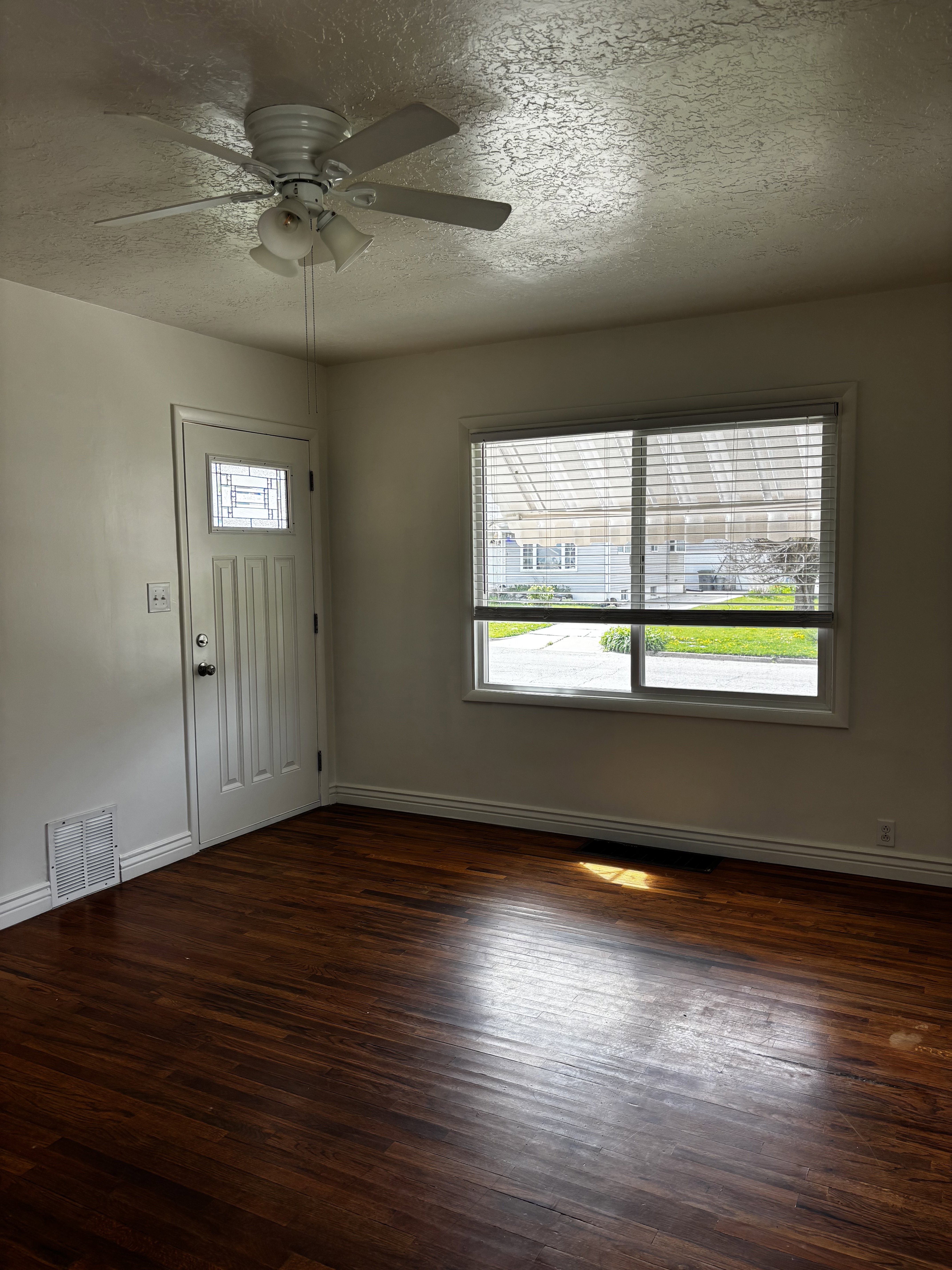 Room featuring rich wood-finish flooring, a white paneled entry door with decorative glass, a window with horizontal blinds, a white ceiling fan with integrated lighting, and white baseboards