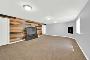 Spacious room featuring a multi-toned wood plank accent wall, wall-mounted fireplace, light gray painted walls, a window with white trim, and a textured carpeted floor
