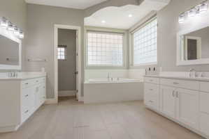 Bright bathroom featuring an expansive soaking tub, dual white vanity cabinets with integrated sinks, and glass block windows