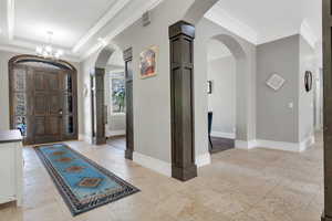 Grand entryway featuring a paneled wood-finish door with decorative glass, tray ceiling with chandelier, arched doorways, wood-finished columns, and light-toned tile flooring