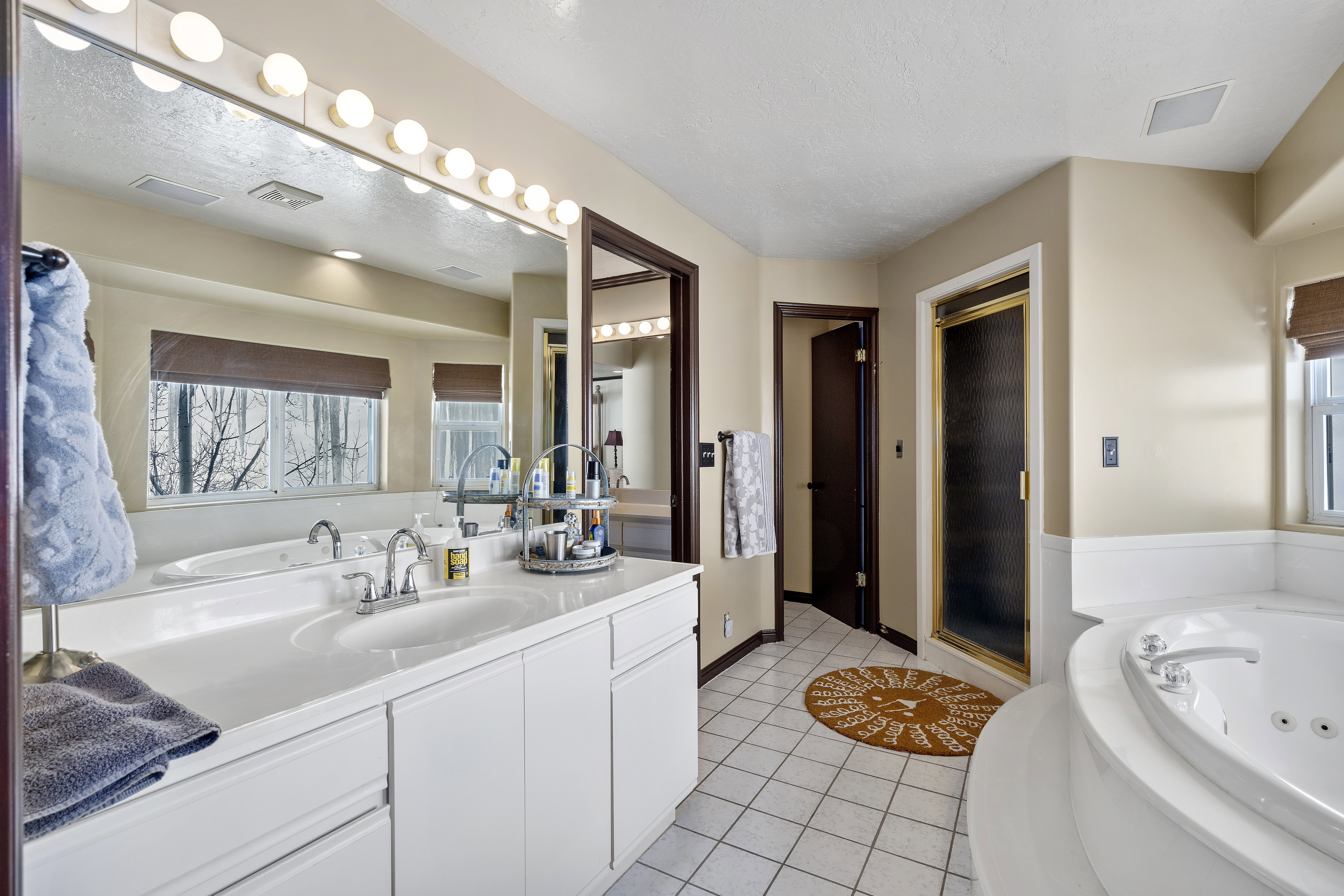 Bathroom featuring a double vanity with integrated sinks, a large mirror with sconce lighting, an enclosed shower with a gold-toned frame, a jetted soaking tub, and white tile flooring