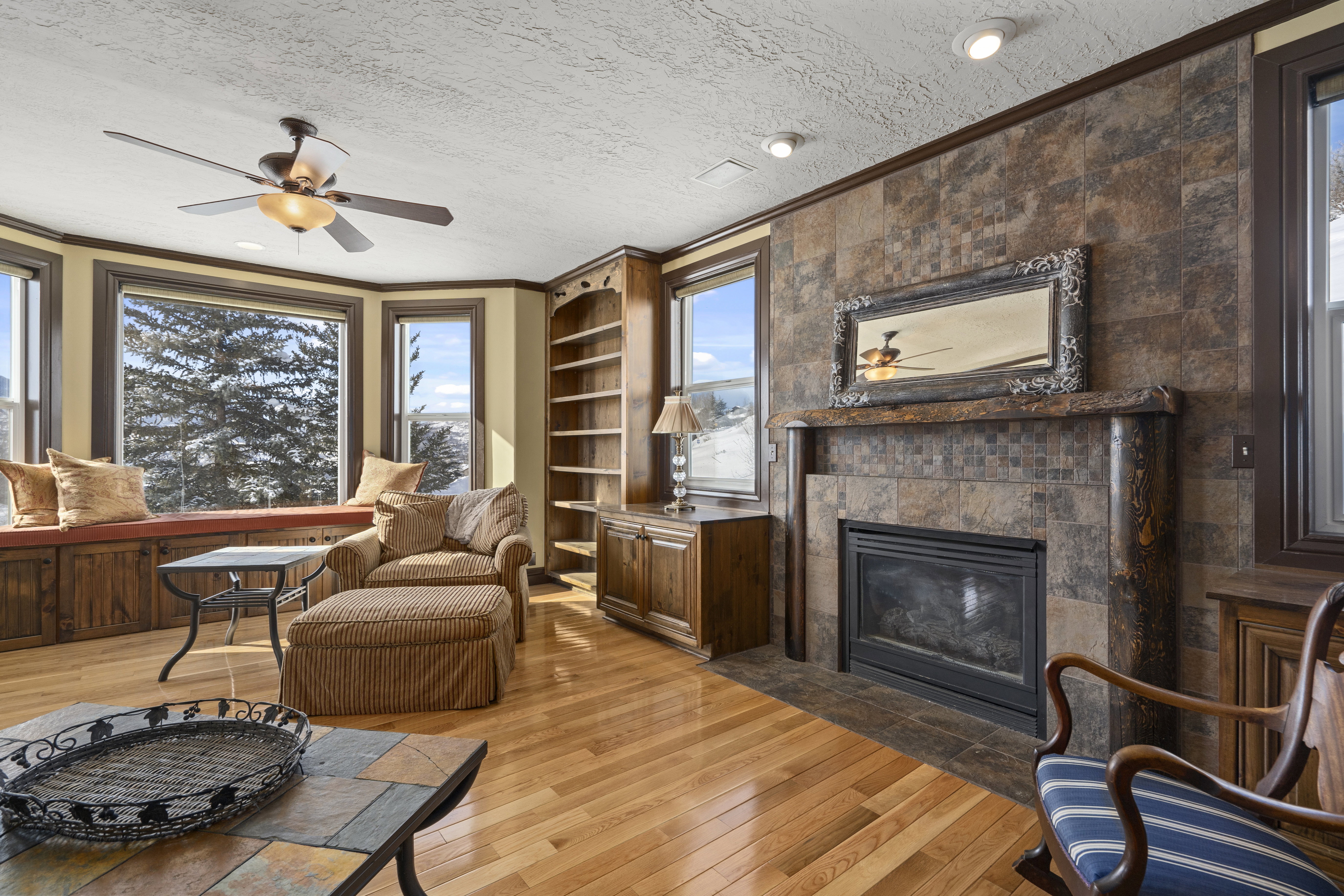 Hardwood flooring throughout a living space featuring a bay window with built-in storage, a ceiling fan, and a fireplace with a stone surround and mantel