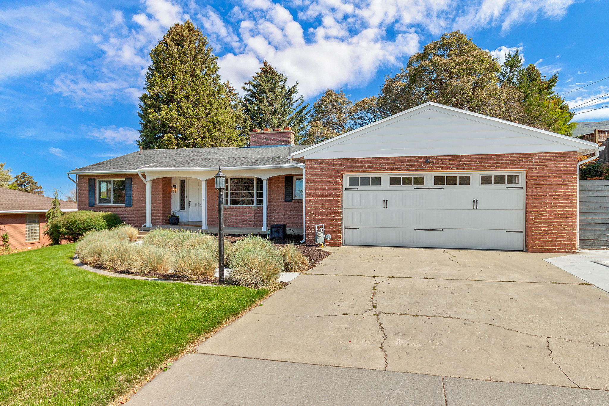 Brick ranch-style exterior featuring a white columned front porch, a two-car garage with white doors and transom windows, and a landscaped front yard with established plantings