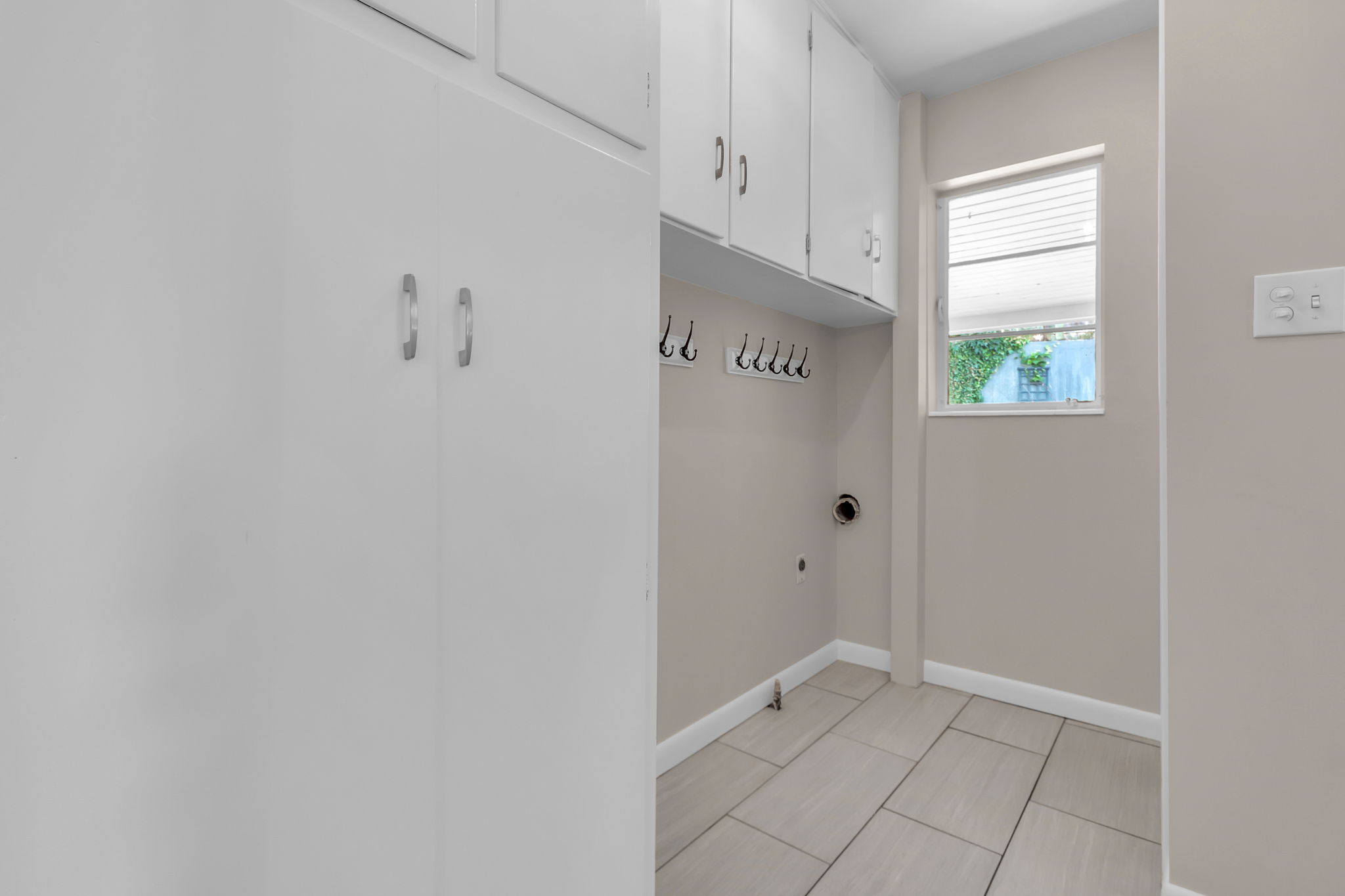 Utility area featuring white upper and lower cabinetry, built-in wall hooks, a window with blinds, and light-toned tile flooring