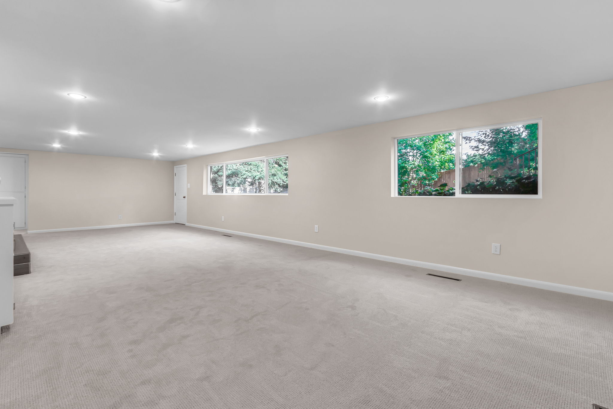 Expansive open room featuring light-toned carpeting, multiple recessed ceiling lights, and two large windows offering greenery outside