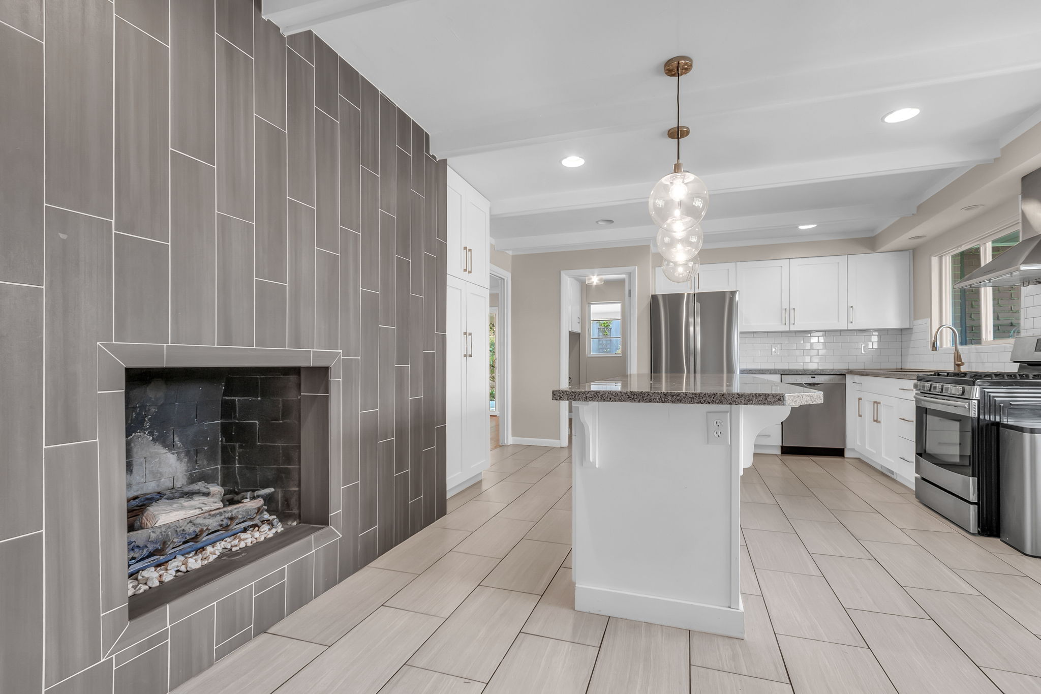 Spacious kitchen and living area featuring a gray tile fireplace, white shaker cabinetry, stainless steel appliances, a kitchen island with seating, and a modern globe pendant light fixture