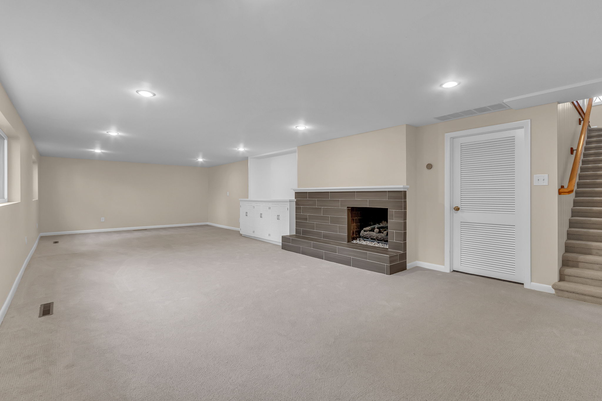 Spacious carpeted room featuring a tiled fireplace, built-in cabinetry, recessed lighting, and a staircase with wood railing