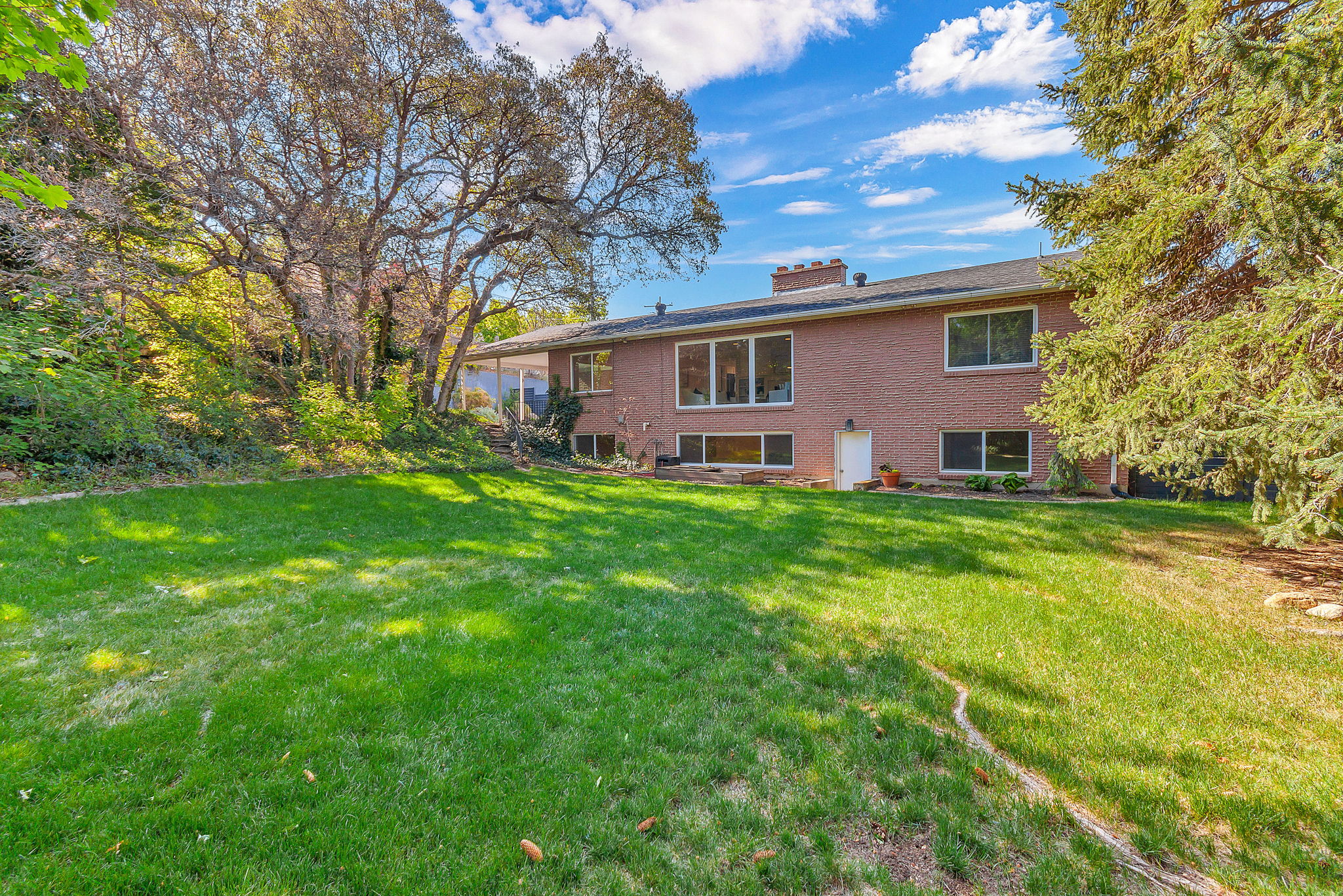 Expansive rear yard featuring a large grass area, mature trees, and a brick exterior home with multiple windows and a covered patio