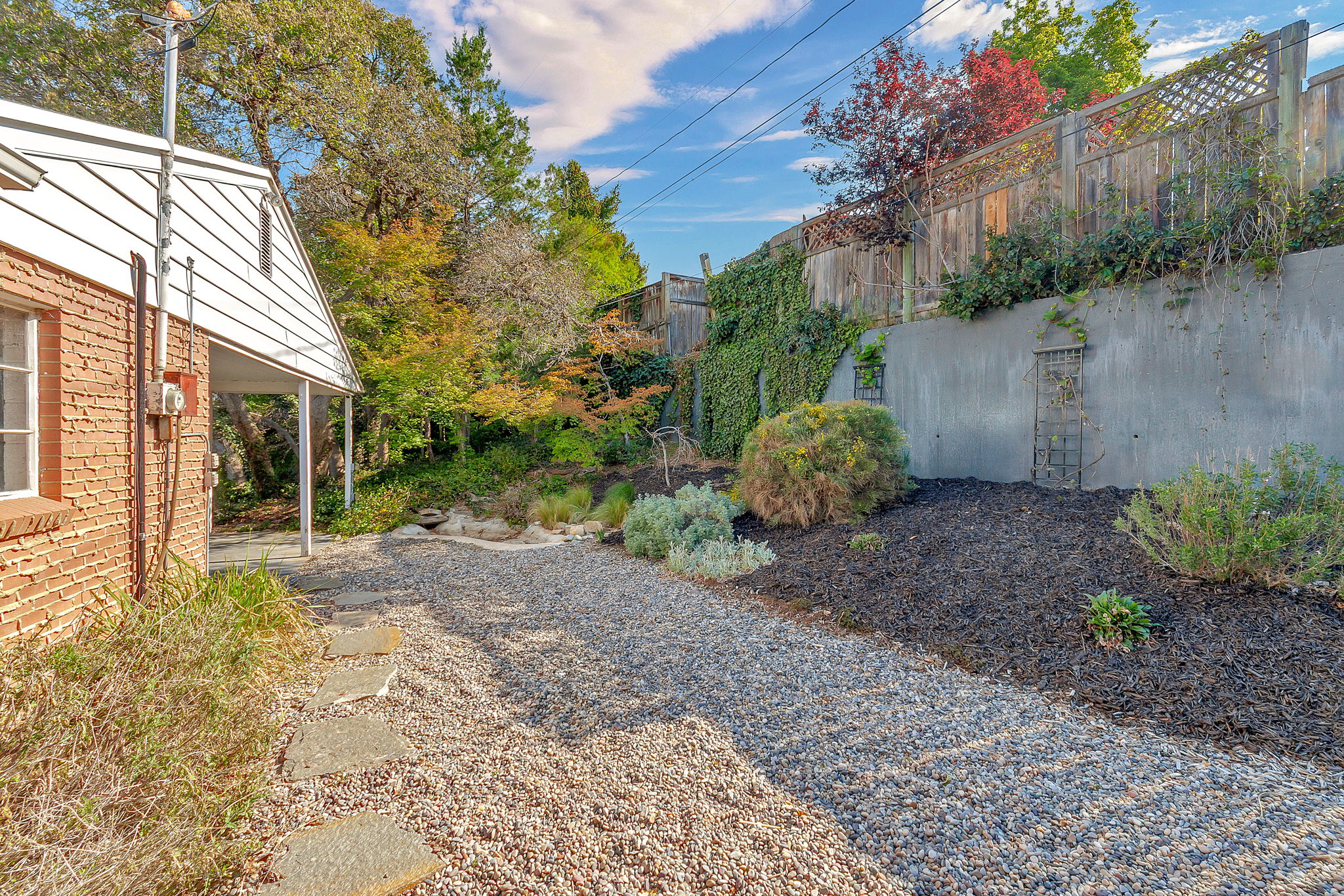 Expansive backyard featuring a gravel pathway with stepping stones, a concrete retaining wall with integrated trellises, a wood privacy fence, and diverse landscaping including mature trees, shrubs, and ground cover
