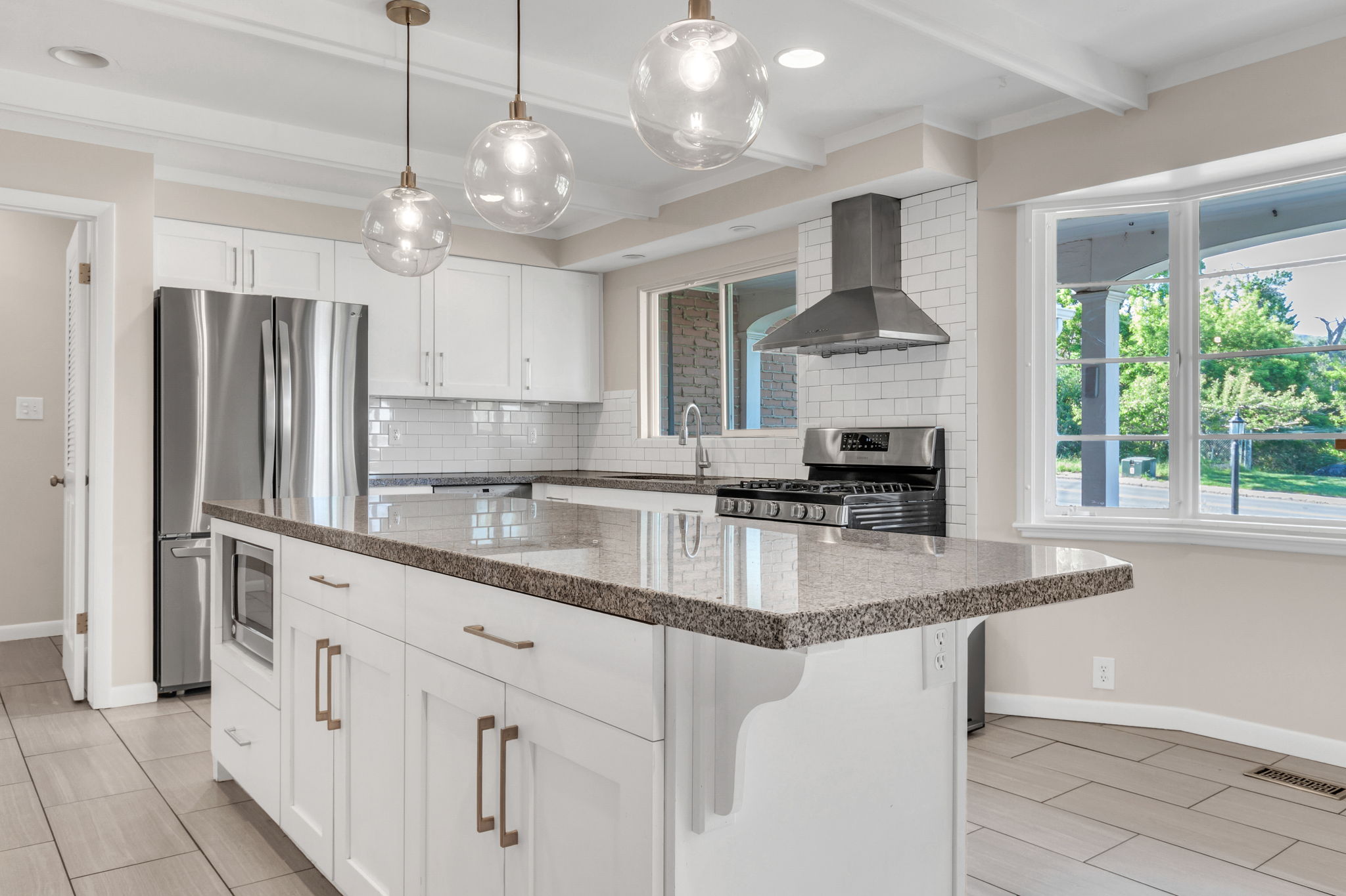 Modern kitchen featuring a large central island with a granite countertop, white shaker cabinetry with bronze hardware, stainless steel appliances, a subway tile backsplash, and decorative pendant lighting