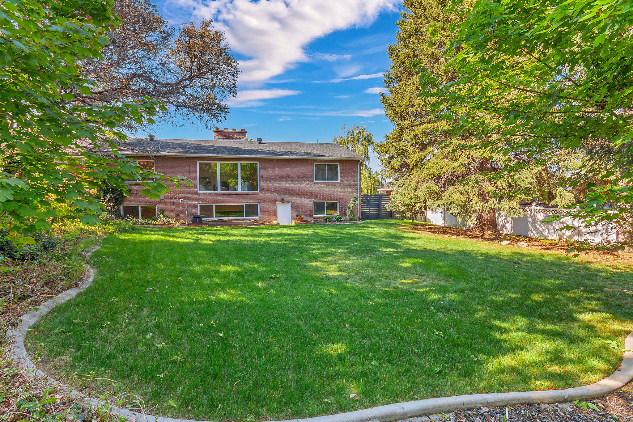 Spacious lawn area framed by mature trees, featuring a brick exterior with multiple windows, a white entry door, and a retaining wall
