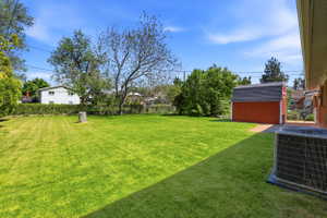 Expansive green lawn featuring a red storage shed with a shingled roof, mature trees, a privacy hedge, and an air conditioning unit