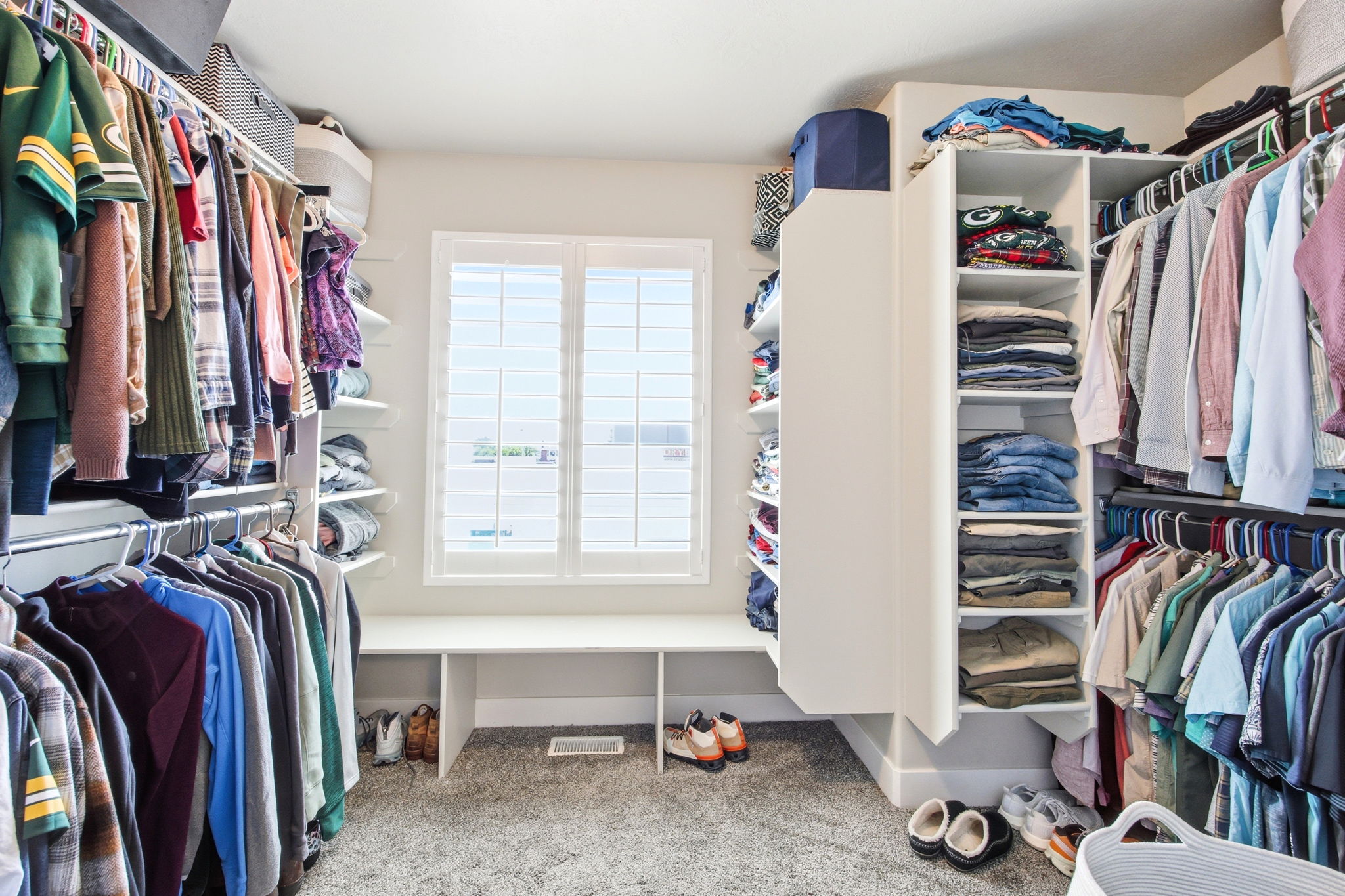 Walk-in closet featuring custom built-in shelving, hanging rods, an integrated bench, and window with plantation shutters