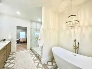 Freestanding soaking tub with floor-mounted brass faucet, walk-in shower with glass enclosure and tile surround, dual vanity with wood-finish cabinetry and light-toned countertop, bead-strand chandelier, and patterned floor tile