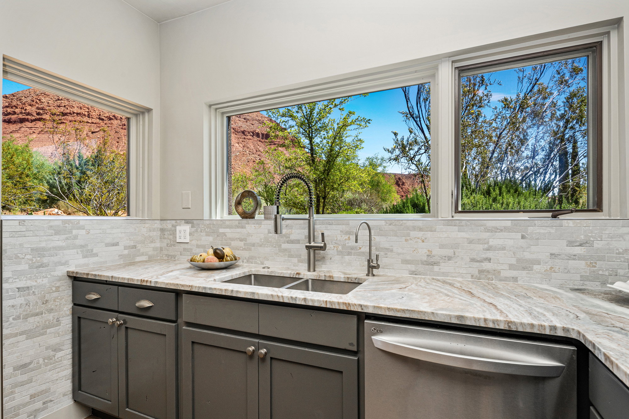Kitchen featuring dual basin stainless steel sink with gooseneck faucet, light-toned stone countertops, and gray shaker-style cabinetry with matte hardware