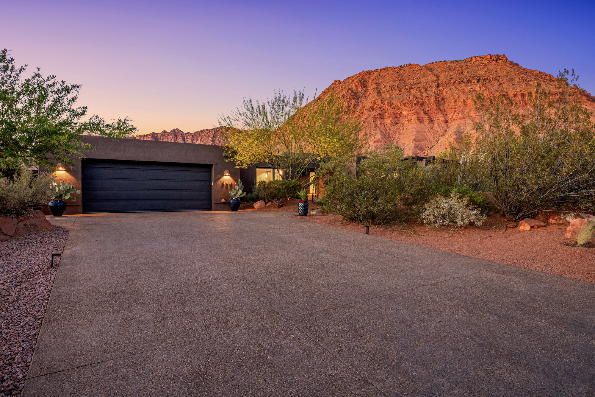 Modern exterior with a double garage door and accent lighting, complemented by desert landscaping and an expansive driveway