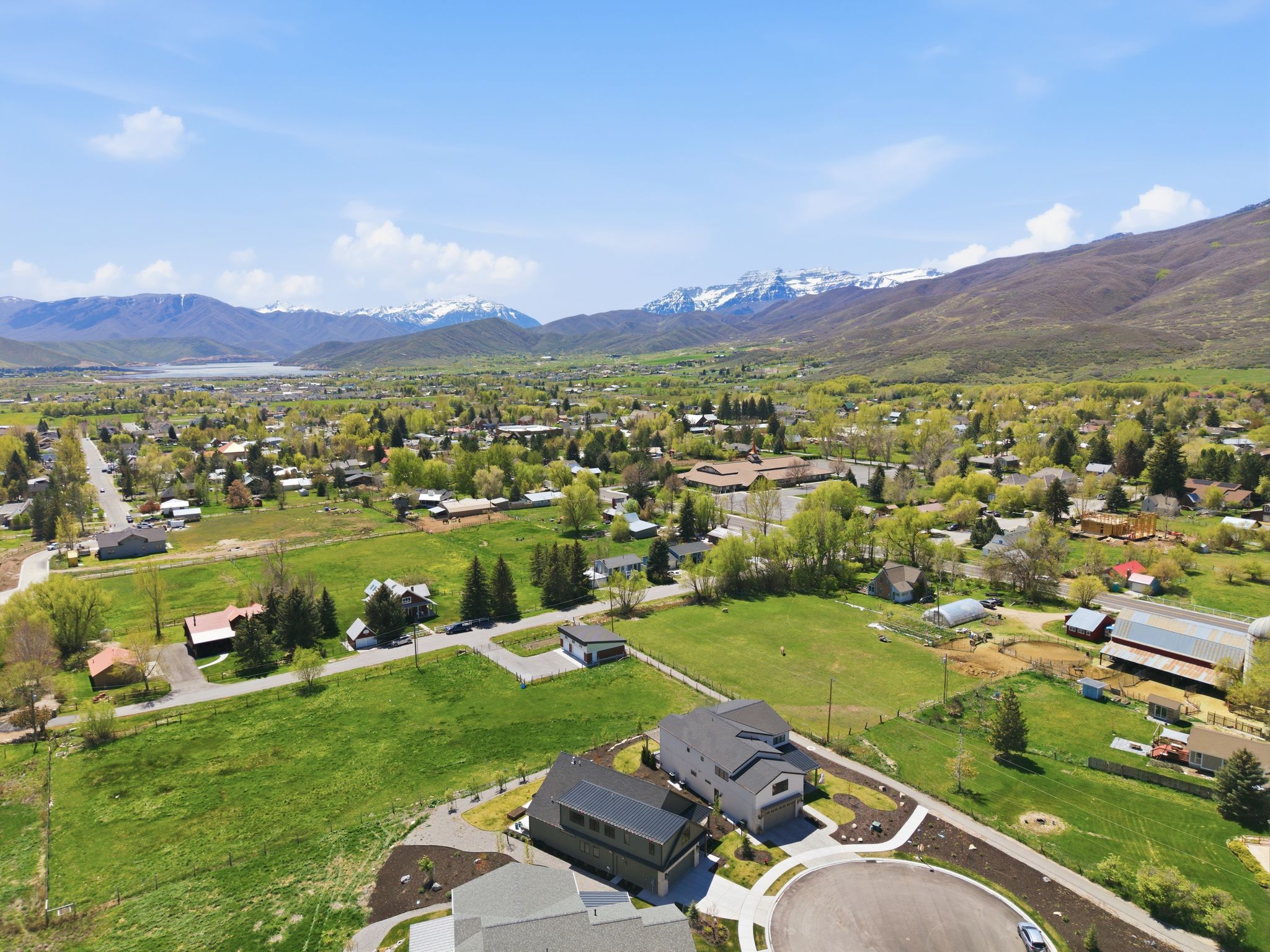 Expansive aerial perspective showcasing mountainous terrain with snow-capped peaks, a distant body of water, and a verdant valley dotted with residential properties