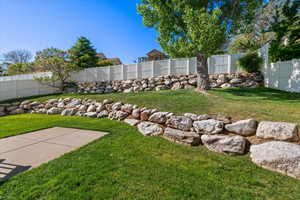 Expansive backyard featuring a concrete patio, verdant lawn, multi-tiered rock retaining walls, mature trees, and privacy fencing
