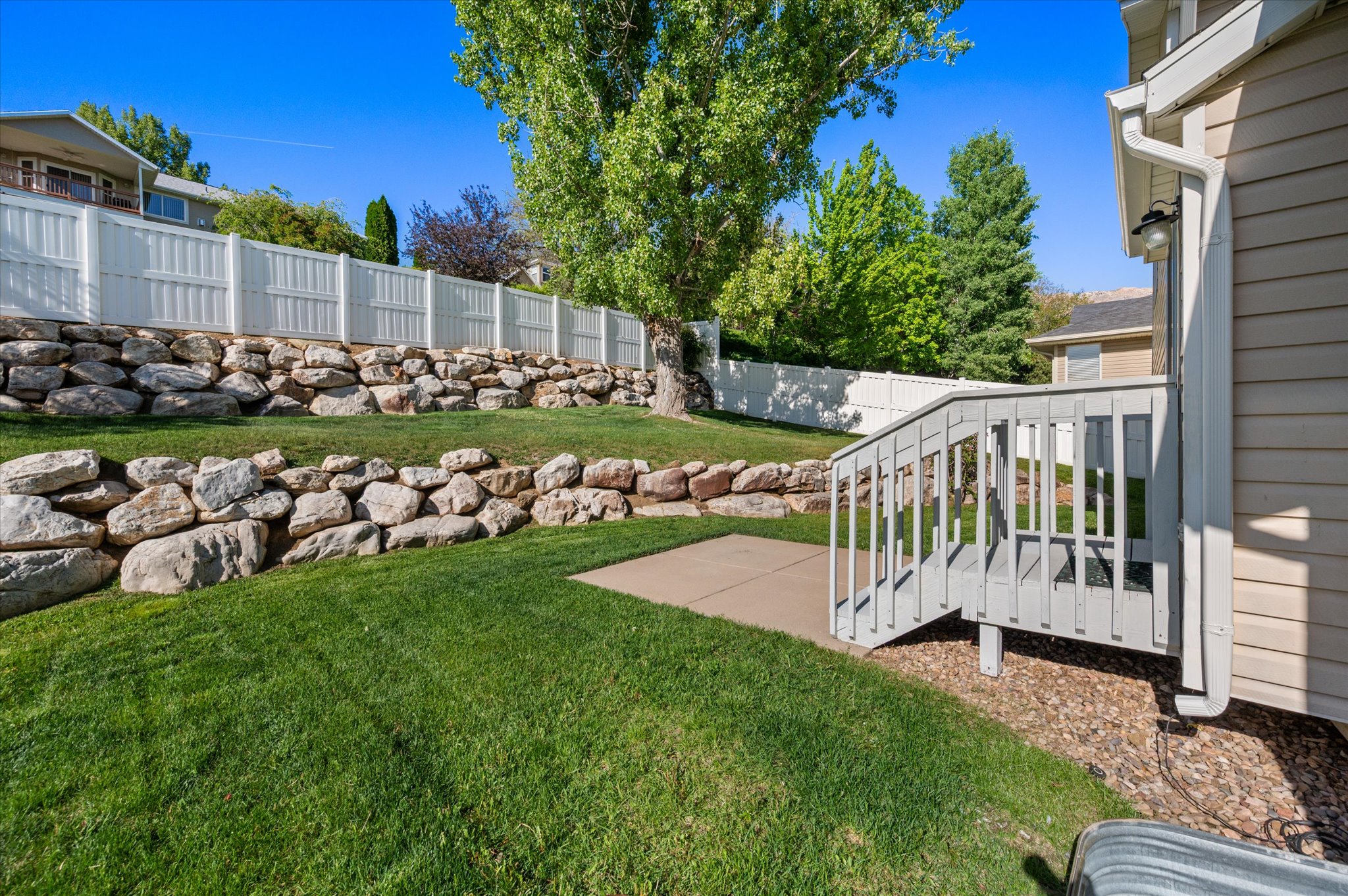 Lawn area featuring multiple natural stone retaining walls, a white vinyl fence, and a wooden deck with stairs