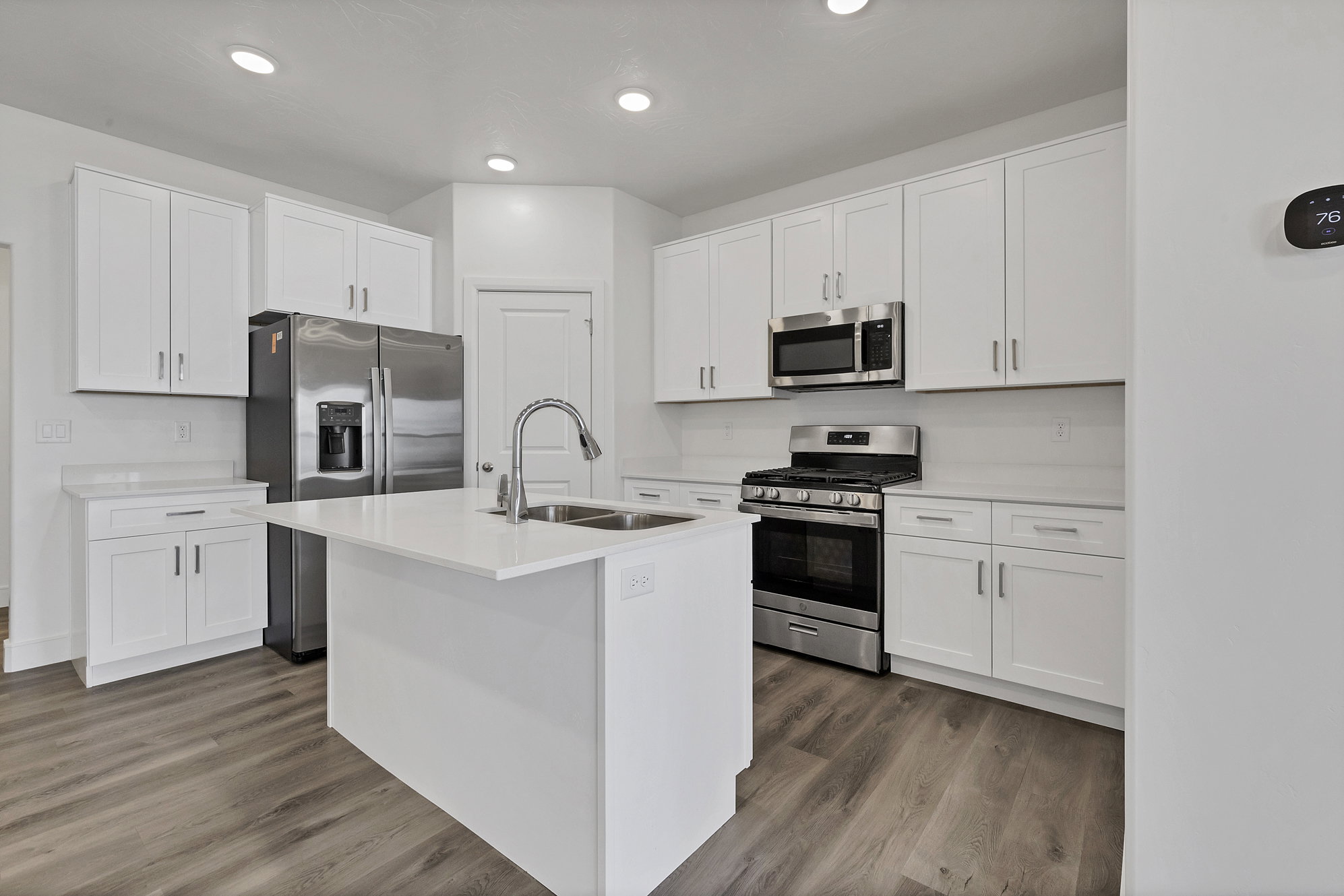 Contemporary kitchen featuring a central island with an undermount sink, white cabinetry with brushed nickel hardware, light-toned countertops, wood-finish flooring, and stainless steel appliances