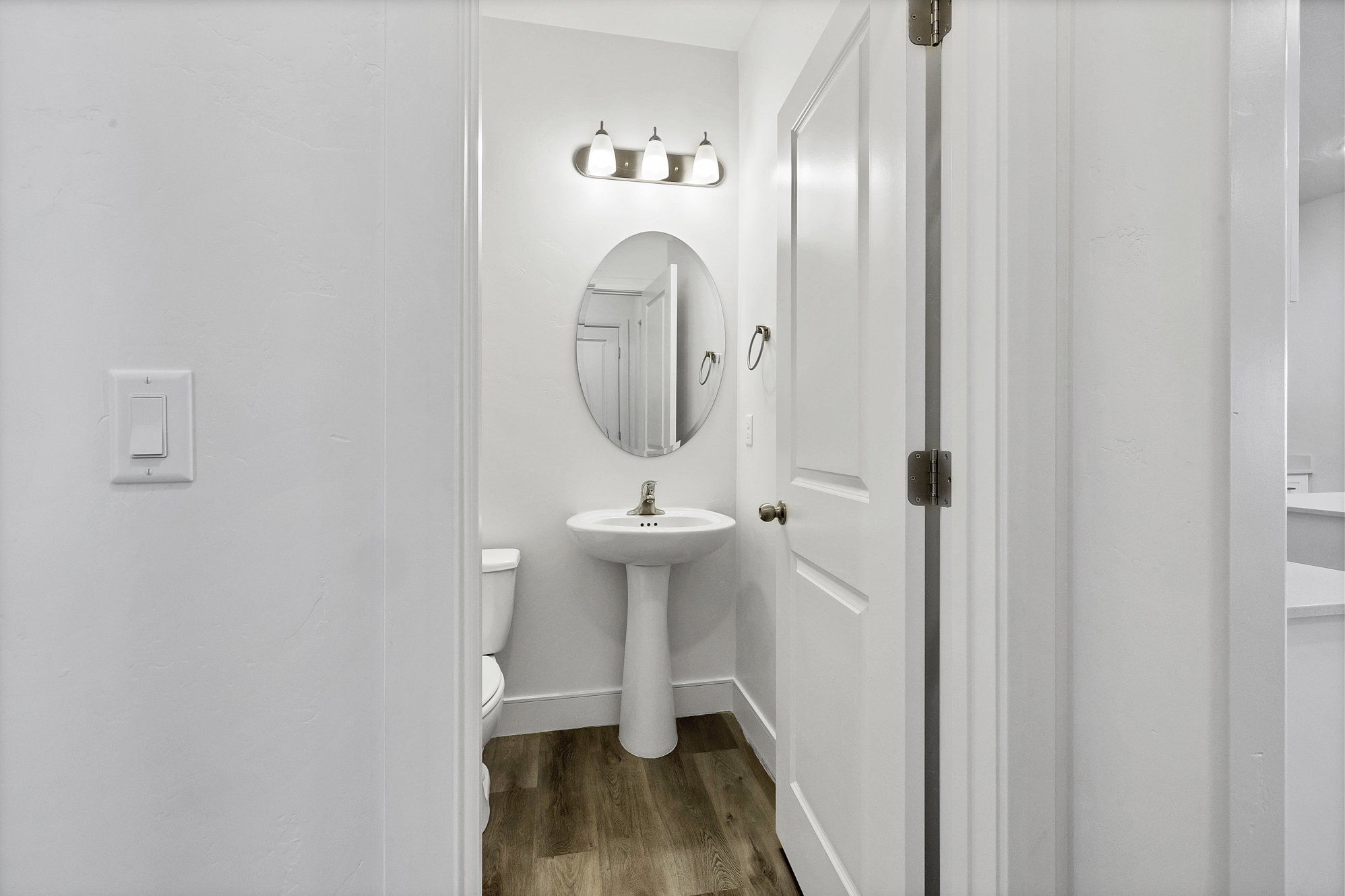 Compact bathroom featuring a white pedestal sink, oval mirror, and wood-finish flooring