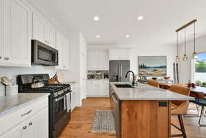 Modern kitchen featuring white shaker cabinetry, stainless steel appliances, light-toned countertops, a central island with a sink, and wood-finish flooring