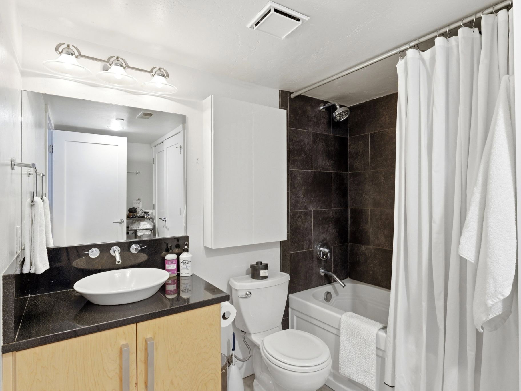Bathroom featuring a vessel sink atop a dark countertop, light wood-finish cabinetry, a built-in tub with dark tile surround, a wall-mounted faucet, and overhead vanity lighting