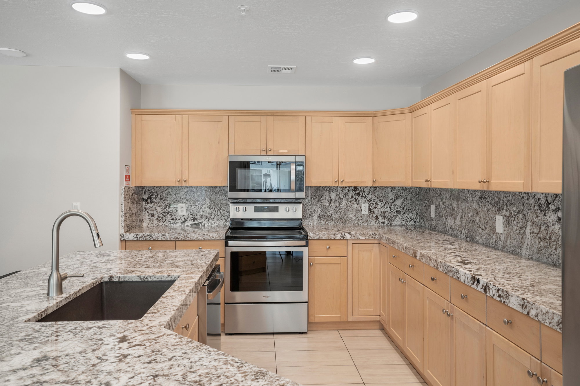 Spacious kitchen featuring light wood cabinetry, stone countertops, a stainless steel sink with a gooseneck faucet, and integrated stainless steel appliances