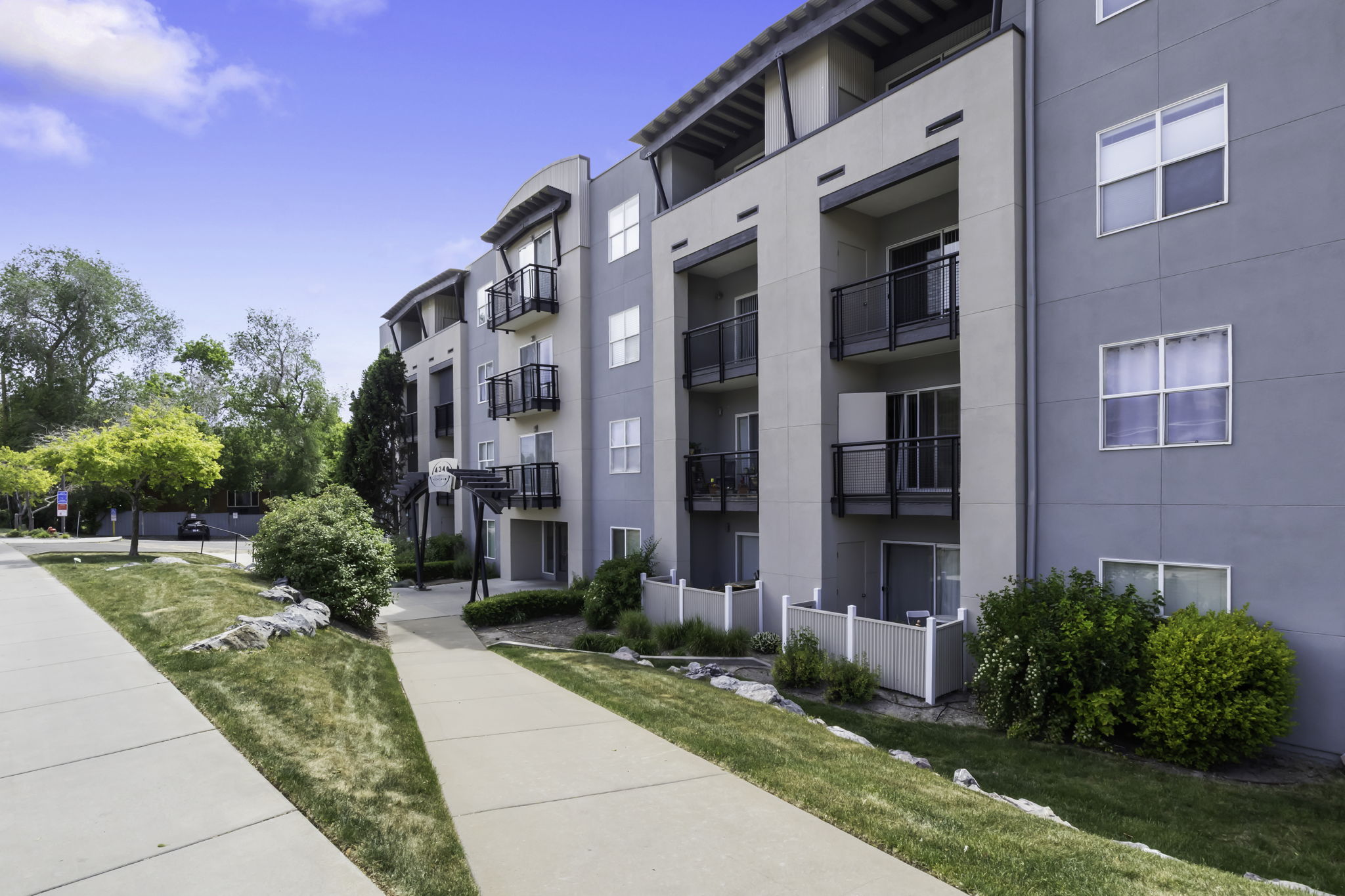 Modern apartment building exterior featuring multiple levels, private balconies with black railings, ground-level patios with white privacy fences, and a pedestrian walkway leading to the entrance